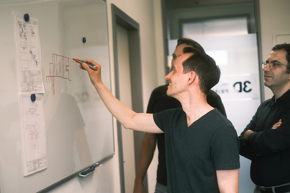 Mitarbeiter der 3D MicroPrint GmbH stehen vor einer Tafel, zeichnen und diskutieren technische Konzepte
